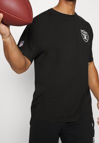 Svart kortärmad T-shirt med rund hals, med en vit Raiders-logotyp på bröstet och en NFL-logotyp på ärmen.
