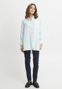 Camicia a maniche lunghe a righe blu chiaro e bianche con taschino frontale e chiusura con bottoni, abbinata a pantaloni blu navy e scarpe nere.