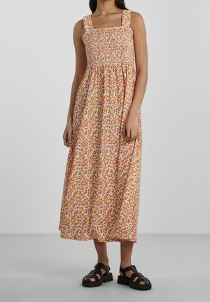 Femme portant une robe midi fleurie smockée sans manches avec de larges bretelles et des sandales noires épaisses, debout devant un fond uni.