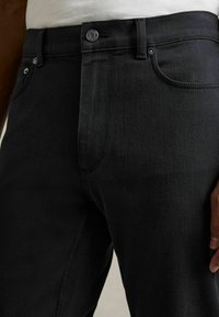Blugi negri din denim cu un design ajustat, care prezintă o buzunare frontală, o închidere cu nasture metalic și cusături subtile în contrast. Textură netedă.