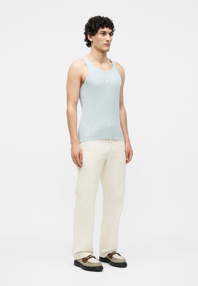 Άνδρας μοντέλο όρθιος, φοράει ανοιχτό μπλε ριμπ φόρεμα χωρίς μανίκια, κρεμ παντελόνι και μπεζ-καφέ loafers σε απλό λευκό φόντο.