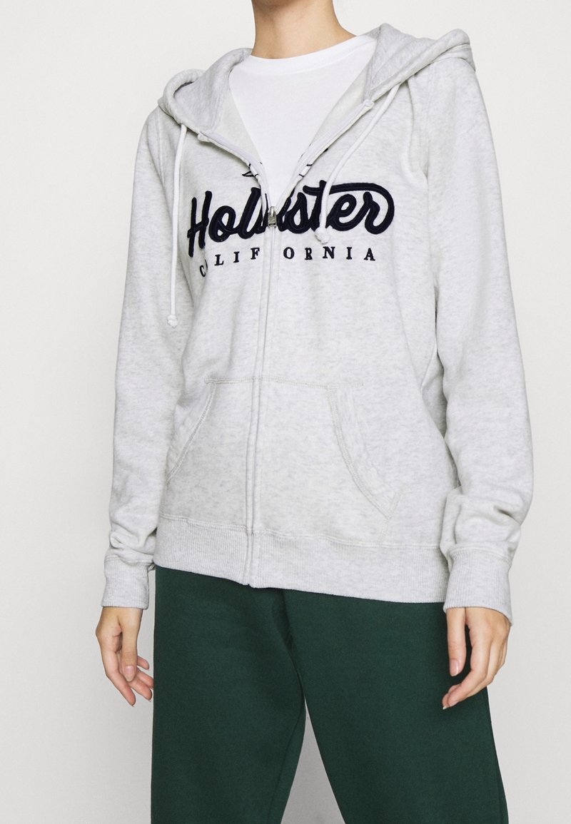 Sudadera gris con cremallera, bolsillo tipo canguro, cordones y logo azul marino. Tela suave, puños y dobladillo acanalados. Combinada con pantalones verde oscuro.
