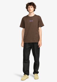Camiseta de algodón marrón con cuello redondo, que presenta un logo en púrpura de "element". Combinada con pantalones de mezclilla negros y zapatos beige.