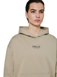 Jong persoon met kort donker haar, die een beige hoodie draagt met de tekst „HALO“ op de borst, kijkt recht vooruit tegen een witte achtergrond.