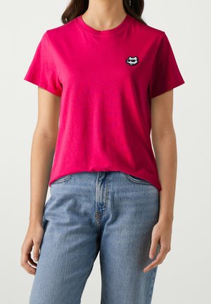 Mujer que lleva una camiseta de manga corta de color rosa brillante con un pequeño parche de gato negro en el pecho, combinada con vaqueros azul claro.