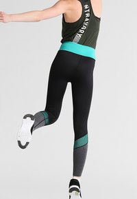 Černé legíny s vysokým pasem a akcenty z modré a šedé síťoviny, v kombinaci s tmavě zeleným sportovním topem s bílou hashtag grafikou.