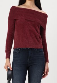 Un pull bordeaux décolleté avec une texture douce et pelucheuse, des manches longues et un détail côtelé au niveau du col et de l'ourlet. Associé à un jean noir.