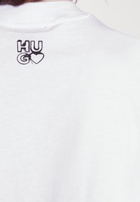 Hvid bomulds t-shirt med en sort grafisk tryk af "HUGO" med et hjerte-motiv på den øverste del af ryggen. Blød tekstur, afslappet pasform.