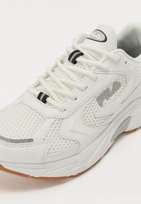Zapatilla deportiva blanca con parte superior de malla y cuero sintético, suela de goma, acentos negros y cordones con lazo. Logo destacado en el lateral.