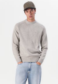 Hellgrau gestrickter Pullover mit einem gerippten Rundhalsausschnitt und Bündchen, kombiniert mit ausgewaschenen blauen Jeans. Eine karierte Kappe rundet den Look ab.
