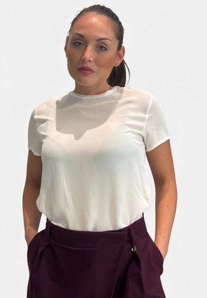 Donna con capelli scuri raccolti indietro che indossa una camicetta bianca trasparente a maniche corte e pantaloni scuri a vita alta con le mani nelle tasche.