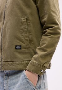 Olive green jacket with zipper, side pocket, and black brand label worn over light blue jeans with hand in pocket.