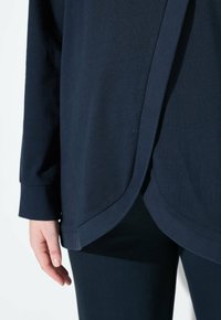 Persona che indossa una maglietta a maniche lunghe blu scuro con orlo arrotondato, sovrapposta a pantaloni blu scuro coordinati, con la mano rilassata al fianco.