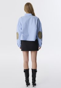 Camicia oversize blu chiaro con toppe a quadri sui gomiti, gonna nera corta e stivali al ginocchio neri con finitura lucida.