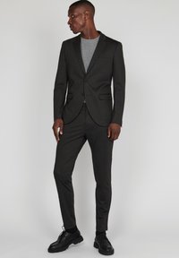 Pikk mees, seljas tumehall slim-fit ülikond ja mustad kingad, seisab ja vaatab vasakule, lihtsa heleda tausta taustal.