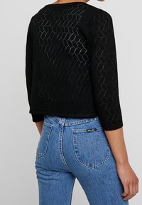 Svart, strukturerad stickad topp med ett honungscomb-mönster, trekvartsärmar och en kort design, tillsammans med blå denimjeans med medelhög midja.