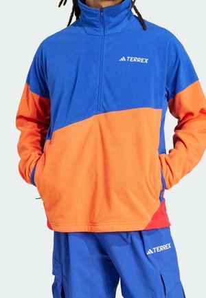 Mies, jolla on sininen ja oranssi Terrex-fleecepusero vetoketjullisella kauluksella ja siniset Terrex-housut, joissa on tavarataskut, kädet puseron taskuissa.