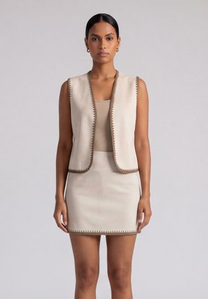 Donna che indossa un gilet beige senza maniche e gonna abbinata con rifinitura marrone decorativa, in piedi davanti a uno sfondo chiaro e neutro.
