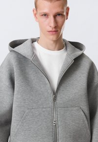 Junger Mann mit kurzen blonden Haaren, der ein weißes Hemd und einen teilweise zugezogenen grauen Kapuzenpullover mit Fronttaschen trägt.