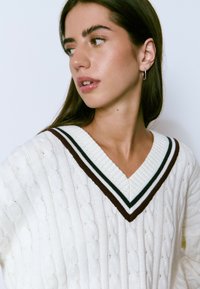 Mujer joven con cabello largo y oscuro que lleva un suéter blanco de punto trenzado con un escote en V a rayas negras y marrones, mirando de lado, arete de argolla plateado visible.