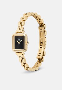 Square gold watch with a black dial, gold link bracelet, and a crown on the side. Features logo "DW" and minimalistic hour markers.