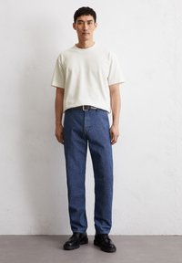 Witte T-shirt met korte mouwen gecombineerd met blauwe denim broek. De broek heeft een rechte snit en een subtiele textuur, aangevuld met zwarte schoenen.