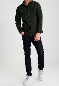 Ciemnozielona koszula z długim rękawem w kamuflażowy wzór; ciemne, dopasowane dżinsy; białe trampki; gładka tekstura; casualowy styl.