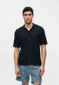 Zwart T-shirt met korte mouwen in een gestructureerde brei met diamantpatroon en een kraag, gecombineerd met blauwe korte broek met patroon. Voorzijde weergegeven.