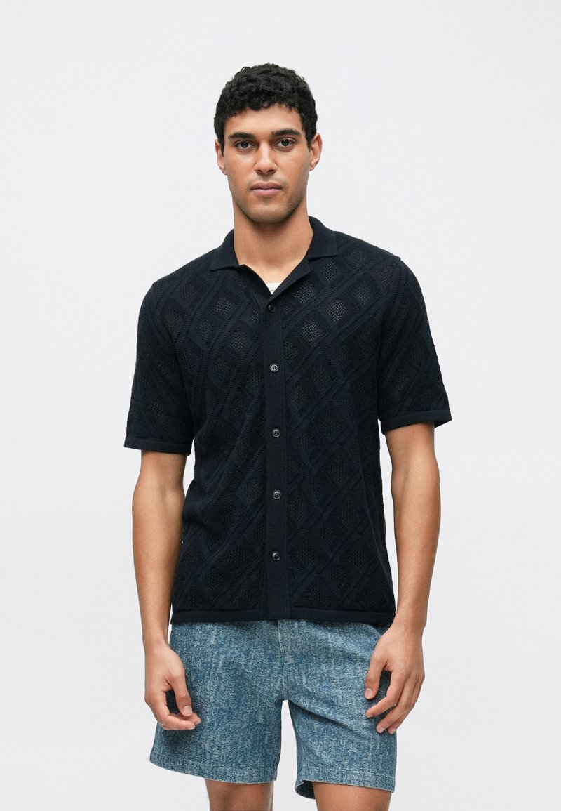 Zwart T-shirt met korte mouwen in een gestructureerde brei met diamantpatroon en een kraag, gecombineerd met blauwe korte broek met patroon. Voorzijde weergegeven.