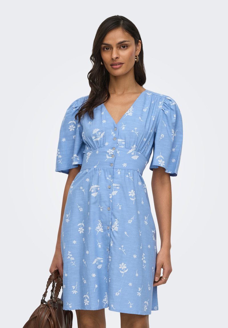 Femme portant une robe bleue à manches courtes avec des motifs floraux blancs, des boutons sur le devant, tenant un sac à main marron tressé.