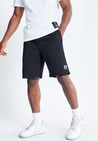 Shorts deportivos negros con una franja lateral blanca, cinturilla elástica y un logotipo impreso. Combinados con zapatillas blancas y calcetines.
