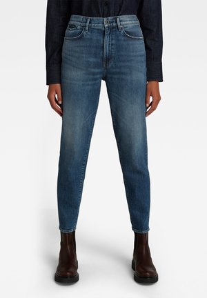 Personne portant un jean en denim bleu taille haute et des bottines marron foncé, debout devant un fond blanc uni.