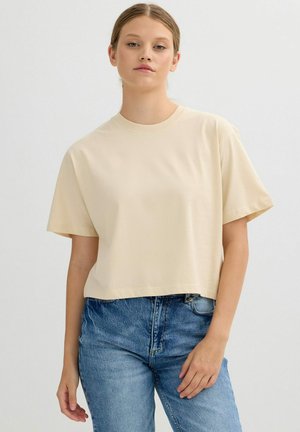 Mujer joven de piel clara y cabello recogido, vestida con una camiseta beige suelta y vaqueros azules, de pie frente a un fondo liso.