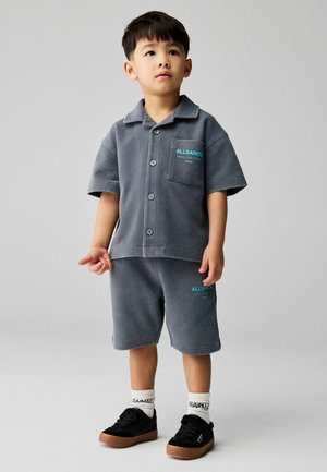 Junge steht und trägt ein graues kurzärmeliges Hemd mit Knöpfen und passende Shorts mit Markenaufschrift, schwarze Turnschuhe und weiße Socken vor grauem Hintergrund.