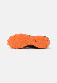 Semelle en caoutchouc orange robuste d'une chaussure de trail running avec des crampons profonds pour l'adhérence, montrée sur un fond clair uni.