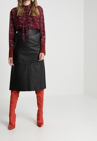 Gonna midi in pelle nera a taglio dritto, abbinata a una blouse floreale bordeaux con colletto a fiocco e polsini a pois rossi, indossata con stivali rossi fino al ginocchio.