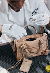 Personne portant des gants blancs inspecte l'intérieur d'un sac à main en cuir beige sur un tapis de découpe noir.