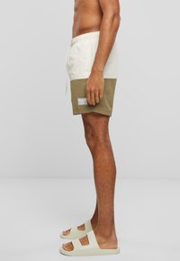 Shorts de plage avec une partie supérieure blanche et une partie inférieure vert olive, taille élastique avec cordon, et un patch logo sur le côté. Portés avec des sandales de couleur claire.