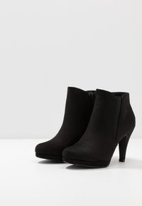 Botas de tobillo de ante negro con tacón alto, punta redonda, cremallera lateral y paneles elásticos para mayor flexibilidad. Textura suave y diseño minimalista.