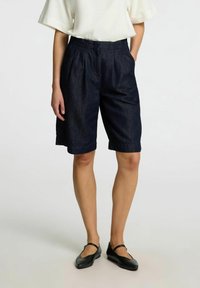 Shorts en denim bleu marine à coupe décontractée, dotés d'une taille élastique et de poches latérales. Portés avec un haut blanc et des ballerines noires.