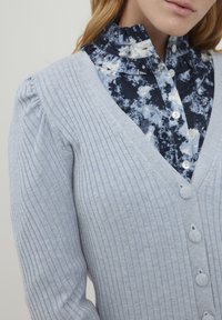 Cardigan côtelé bleu clair avec un col en V, doté de quatre boutons ronds. En dessous, une chemise à fleurs sombres avec un col à volants.