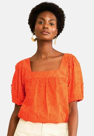 Kobieta z krótkimi kręconymi włosami, nosząca duże złote kolczyki koła i pomarańczowy teksturowany top z bufkami i kwadratowym dekoltem.