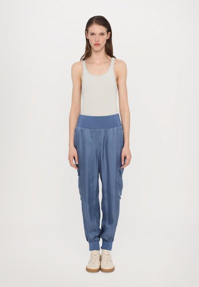 Lichtgrijze tanktop gecombineerd met blauwe, tapered cargobroek met elastische tailleband en zijzakken, aangevuld met crèmekleurige schoenen.