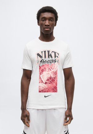 Jaunas vyras dėvi baltą „Nike Hoops“ marškinėlį su raudonu krepšinio grafiku ir baltas „Nike“ šortus, stovintis prieš baltą foną.