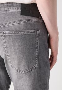 Pilki džinsai su užpakaliniu kišenėliu ir juodu Calvin Klein etiketė. Audinys turi lygų paviršių su matomais siuvinėjimo detalėmis.