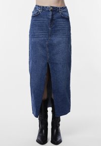 Farmernadrág szoknya elöl hasítékkal, magasan ülepelt, kék színű, texturált anyagból készült, látható varrásokkal. Fekete cowboy csizmával viselve.