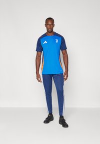 Camisola desportiva azul com detalhes laranja e logótipo, combinada com calças azuis com riscas brancas laterais. Feita de tecido respirável.