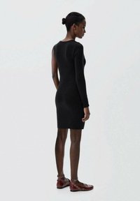 Robe en tricot noir côtelé à manches longues, forme ajustée, encolure ronde et ourlet au-dessus du genou, portée avec des chaussures plates rouges.