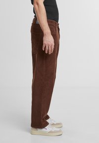 Brown corduroy pants featuring a straight cut, five pockets, and a textured ribbed pattern, worn with light-colored sneakers.