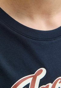 T-shirt in cotone blu navy con collo rotondo e collare a costine; presenta un design stampato in bianco e marrone vicino al fondo. Tessuto testurizzato.